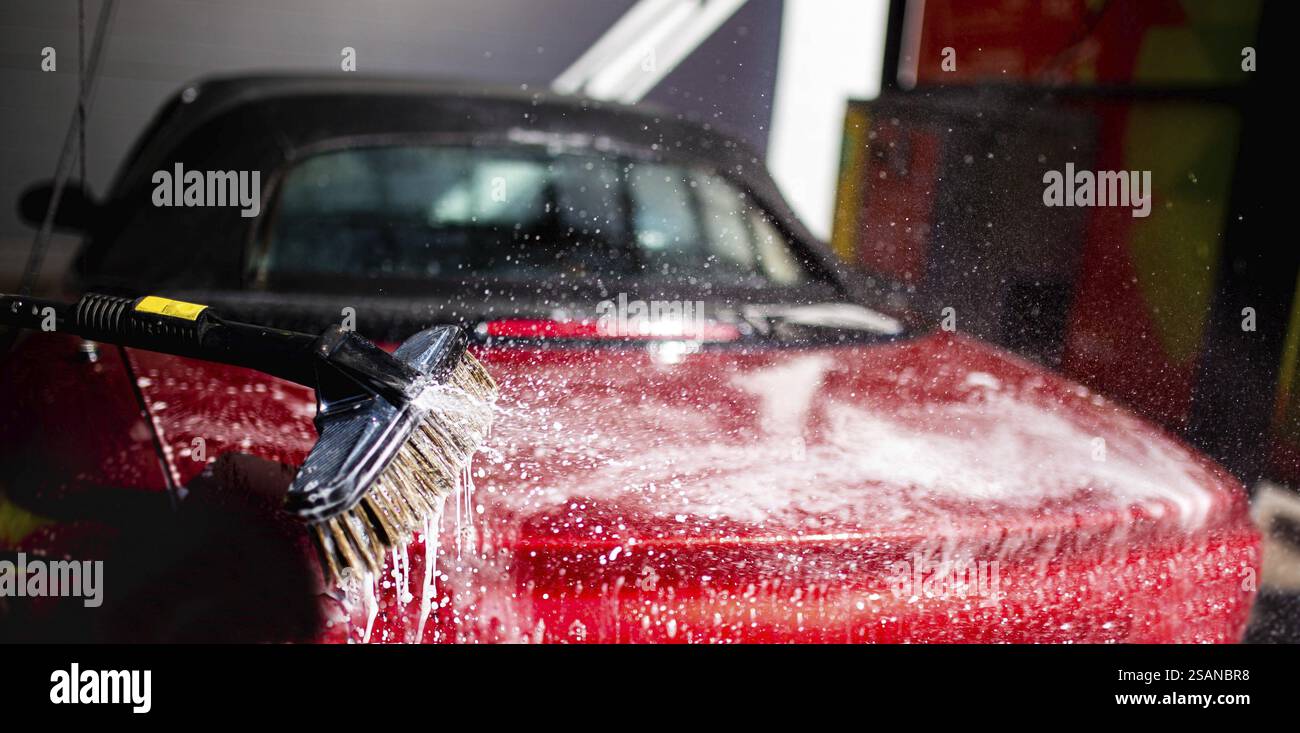Eau savonneuse éclaboussant sur une voiture rouge tout en lavant avec une brosse Banque D'Images