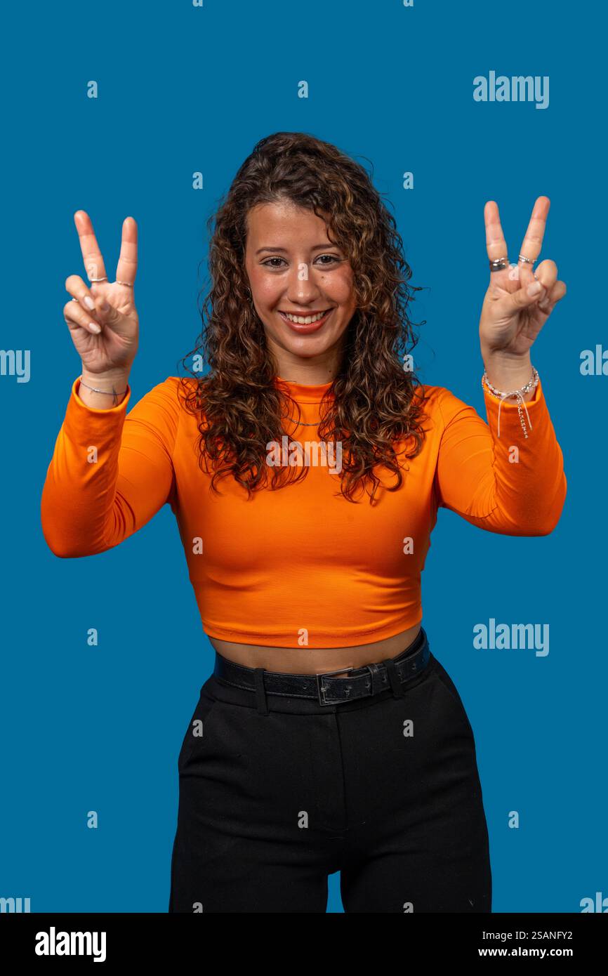 Portrait studio d'une jeune femme souriante faisant signe de paix avec les deux mains sur un fond bleu Banque D'Images