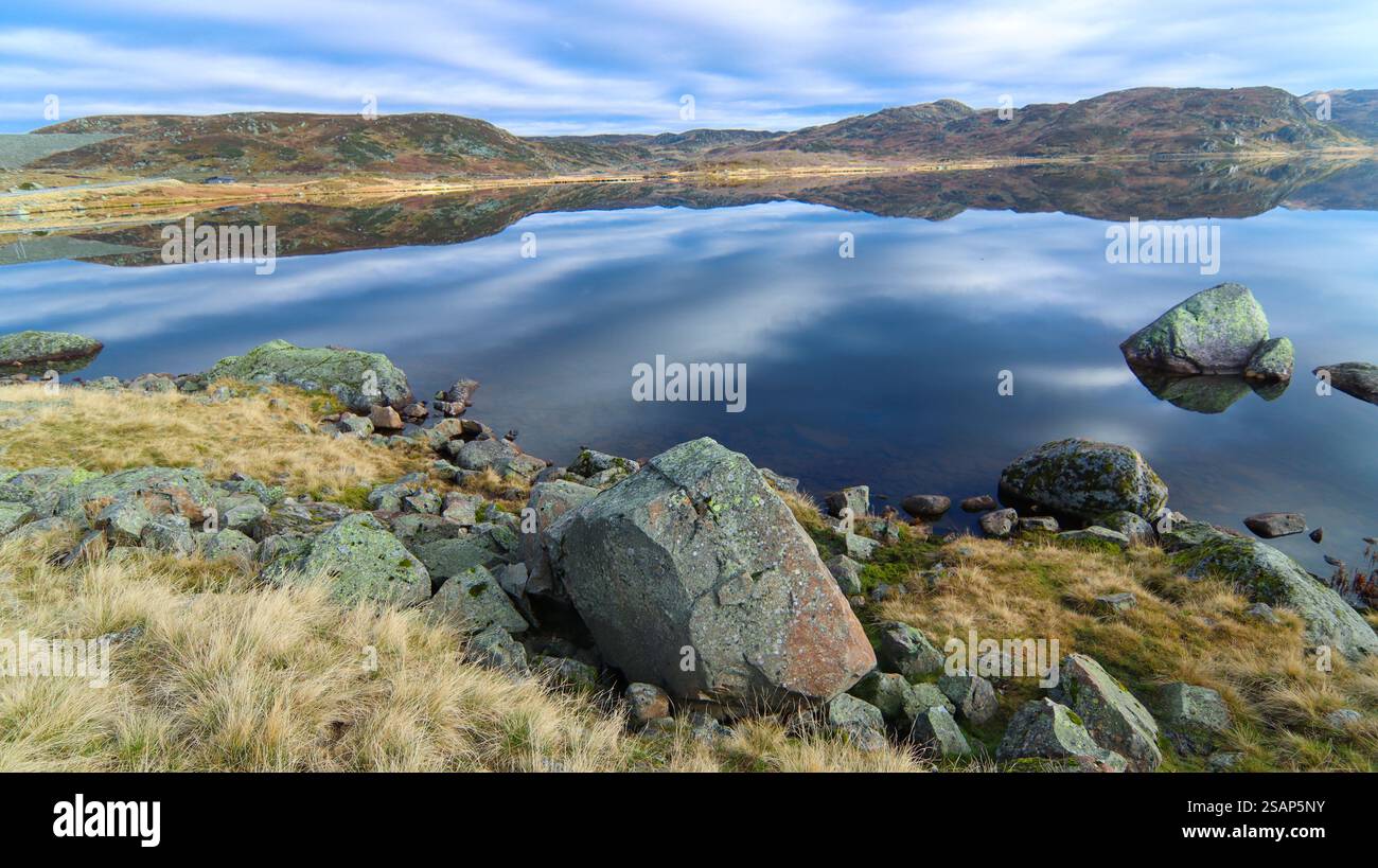 Paysage serein avec des eaux calmes reflétant un ciel nuageux sur un lac de la rive rocheuse pendant la journée. Norvège Banque D'Images Paysage serein avec des eaux calmes reflétant un ciel nuageux sur un lac de la rive rocheuse pendant la journée. Norvège Banque D'Images