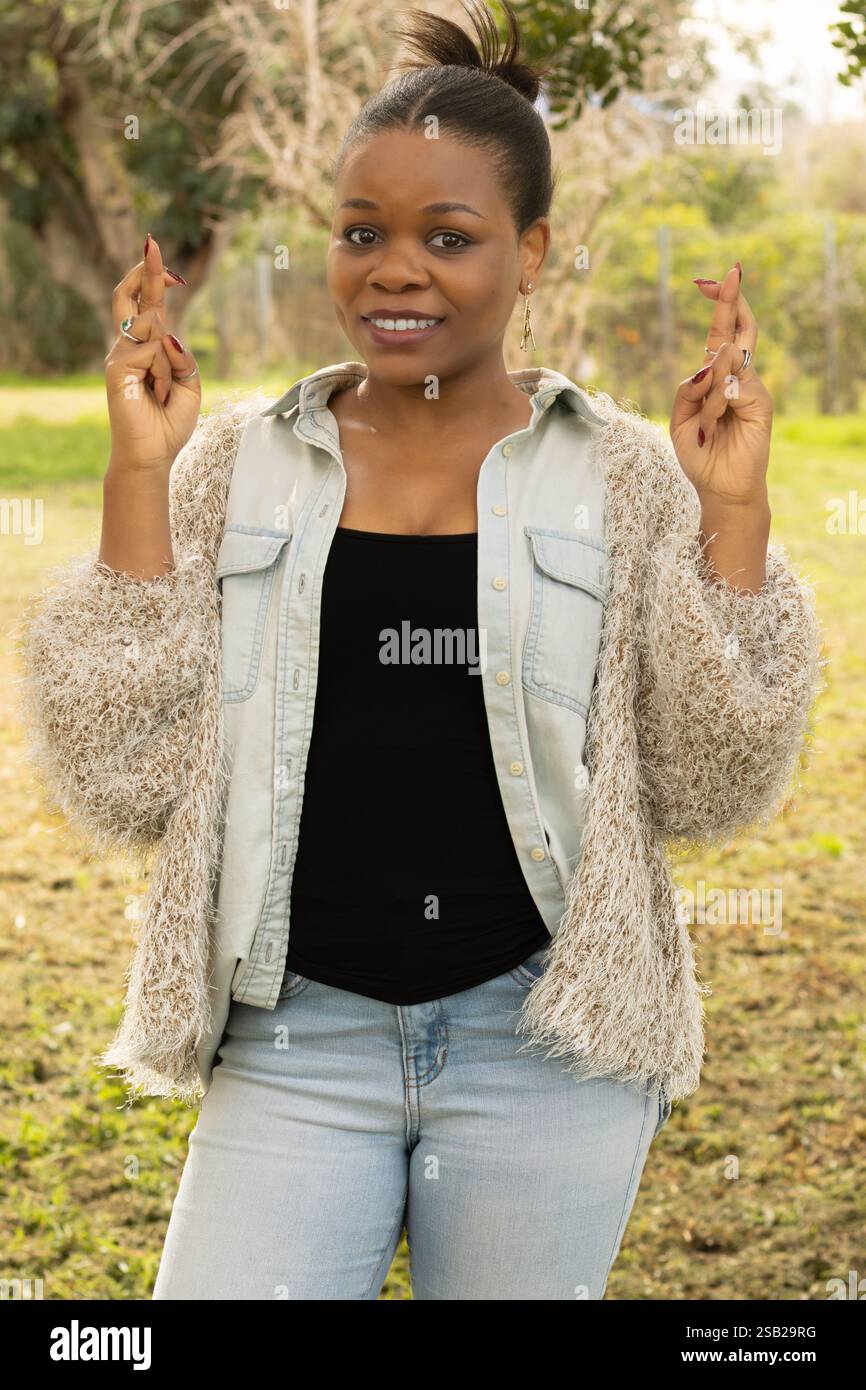 Une femme noire se tient dehors avec une expression joyeuse, croisant les doigts sur les deux mains comme symbole d'espoir et de bonne chance. Elle porte un de élégant Banque D'Images