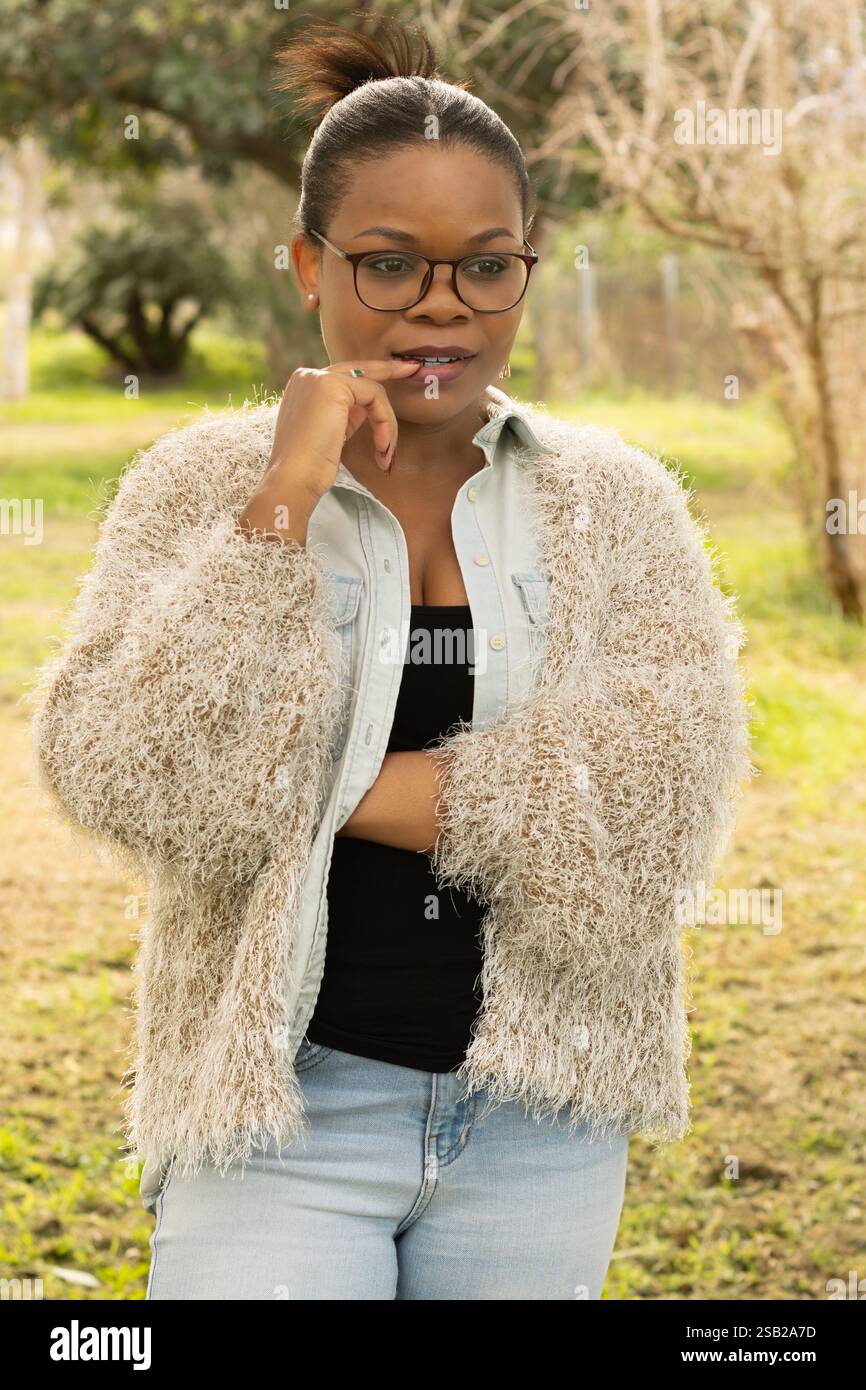 Une femme noire portant des lunettes et une tenue confortable se mord la lèvre tout en regardant au loin avec une expression réfléchie. Banque D'Images
