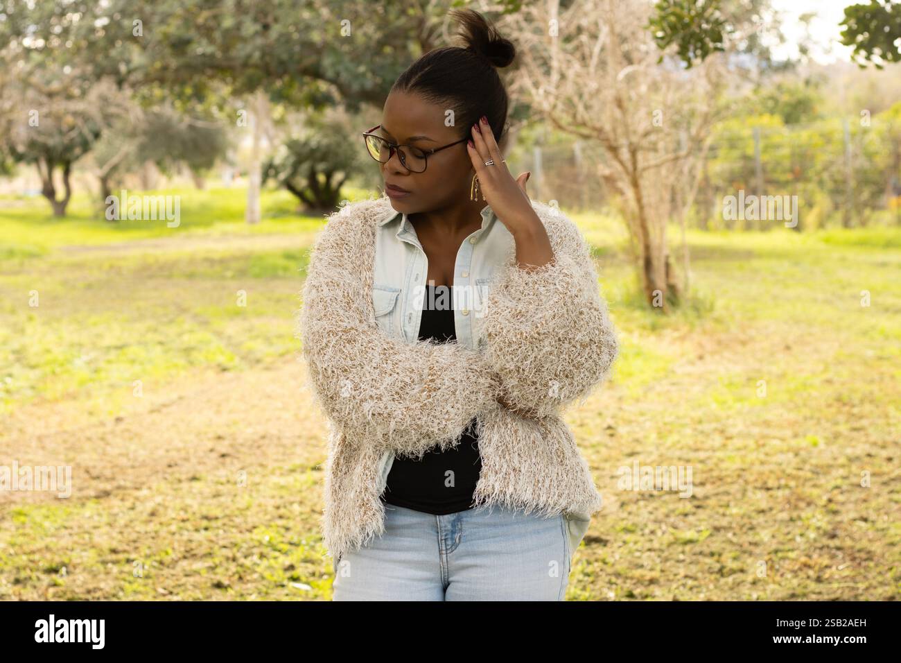 Une femme noire portant des lunettes et un pull beige confortable se tient à l'extérieur, apparaissant profondément dans la pensée. Sa main touche sa tête, et elle regarde vers le bas Banque D'Images