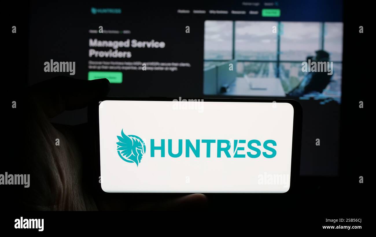 Dans cette illustration photo, une personne tient un téléphone portable avec le logo de la société américaine de cybersécurité Huntress Labs Inc sur l'écran devant la page Web de l'entreprise. Banque D'Images