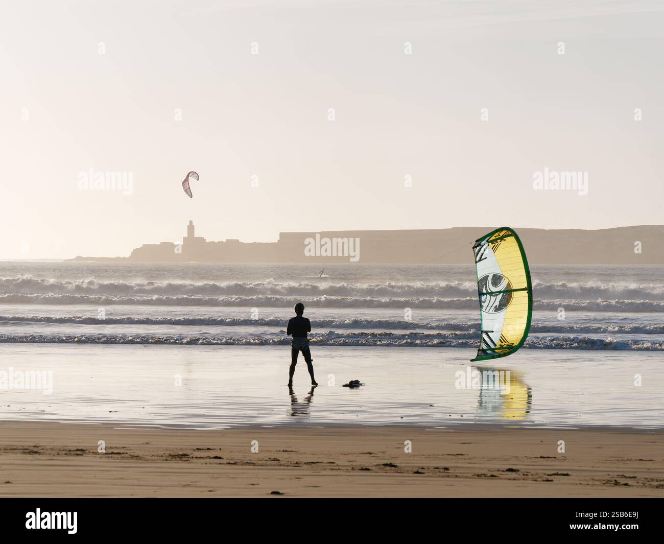 Cerf-volant en silhouette avec cerf-volant jaune se tient sur une plage de sable avec une île derrière dans la ville d'Essaouira, 27-29, 2025 Banque D'Images