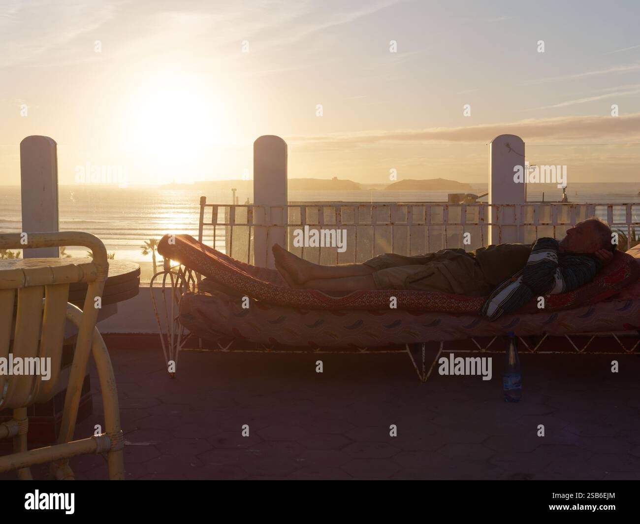 Quelqu'un est allongé sur un transat sur une terrasse alors que le soleil se couche sur la mer une île derrière dans la ville d'Essaouira, 27-29, janvier 2025 Banque D'Images