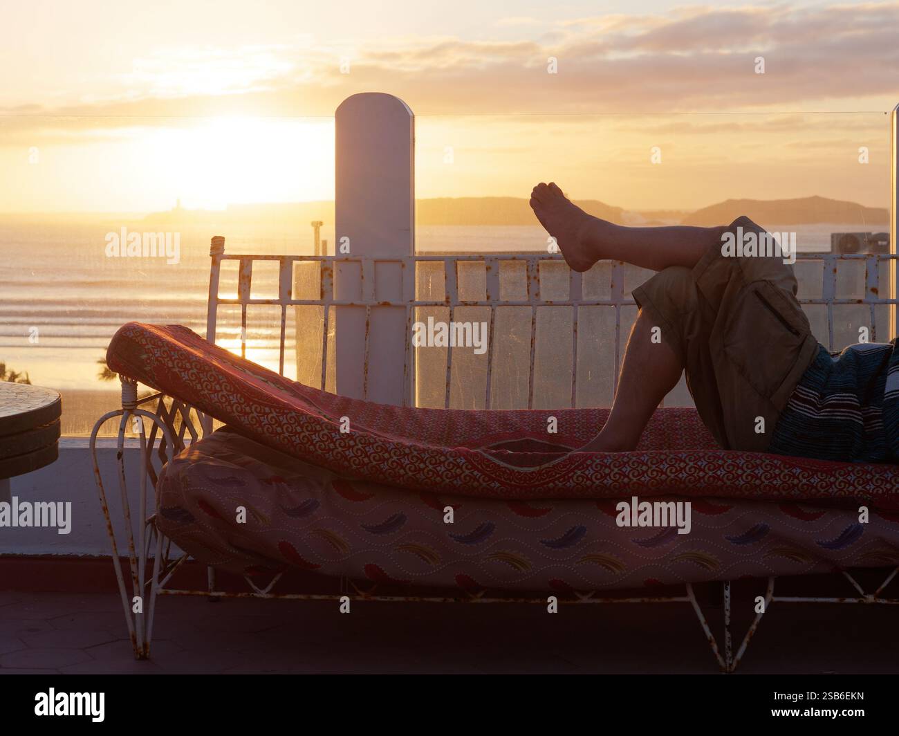 Quelqu'un est allongé sur un transat sur une terrasse alors que le soleil se couche sur la mer une île derrière dans la ville d'Essaouira, 27-29, janvier 2025 Banque D'Images