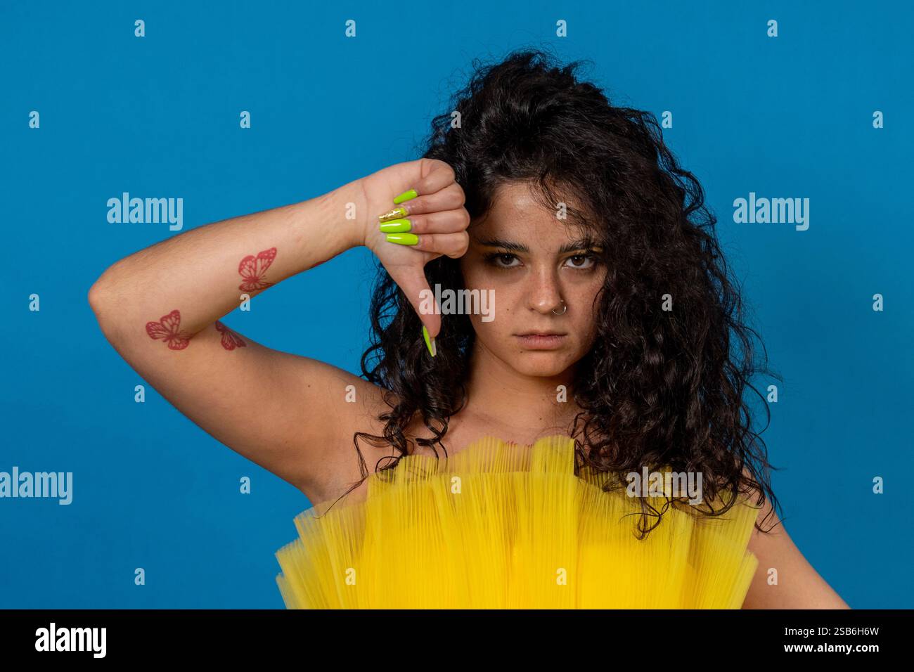 Jeune femme avec des cheveux bouclés et des tatouages papillon montrant les pouces vers le bas, exprimant sa désapprobation et des commentaires négatifs Banque D'Images