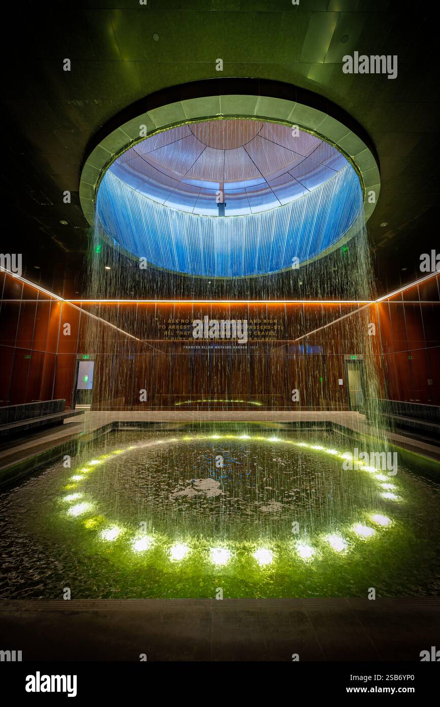 Fontaine de la cour contemplative du Musée national d'histoire afro-américaine à Washington DC | WASHINGTON D.C., États-Unis — illuminée par le bas, la fontaine de la cour contemplative envoie une nappe d'eau à travers un oculus brillant, l'équivalent souterrain du Musée national d'histoire et de culture afro-américaine au Washington Monument qui s'élève juste à l'extérieur. La cour occupe le niveau 0 du hall, le plus profond des étages publics du musée, dans un bâtiment où environ 60 % de la structure se trouve sous le National Mall. Banque D'Images