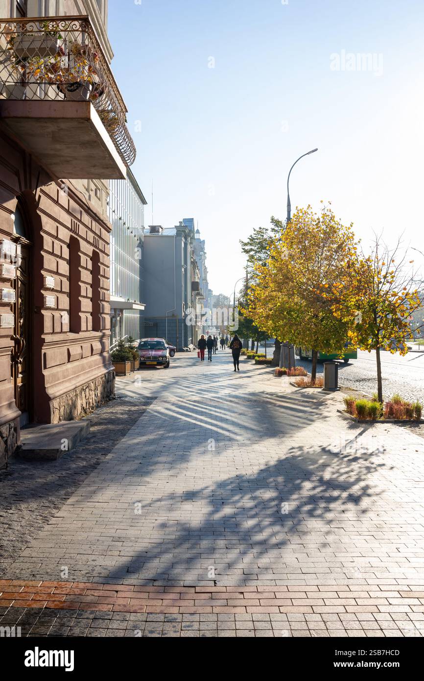 Une passerelle urbaine paisible présente des feuilles d'automne colorées sous un ciel bleu clair. Les piétons se promènent le long du chemin à côté de charmants bâtiments, créant Banque D'Images
