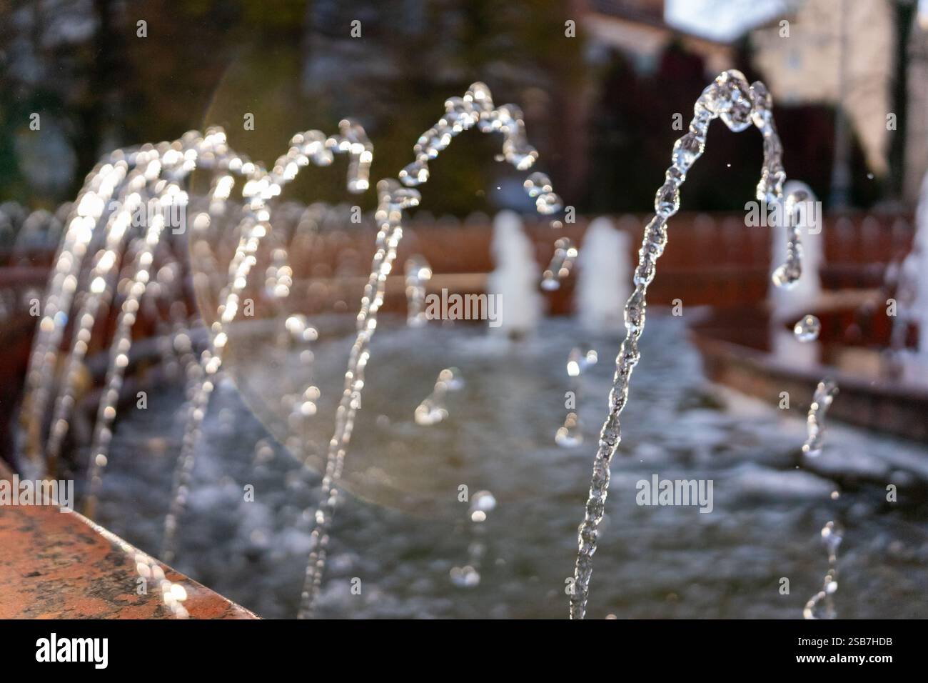 Les cours d'eau sortent gracieusement de la fontaine, scintillant à la lumière du soleil. Les plantes et les arbres environnants créent une atmosphère sereine, invitant les visiteurs à Banque D'Images