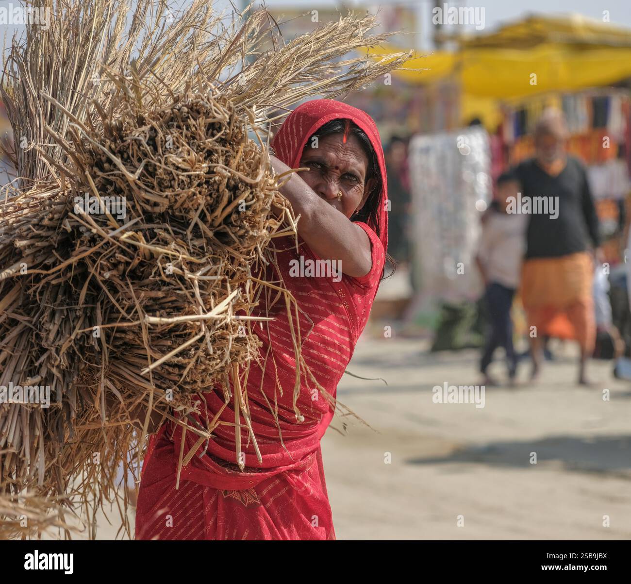 Ouvrier rural vêtu de vêtements rouges vifs transportant du blé récolté pendant la Maha Kumbh Mela à Prayagraj. Le marché animé met en valeur la tradition Banque D'Images