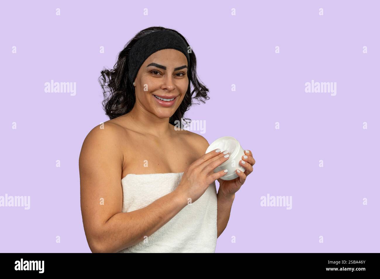 Femme heureuse portant un bandeau et une serviette tenant un pot de crème hydratante, favorisant les soins de la peau et les produits de beauté Banque D'Images