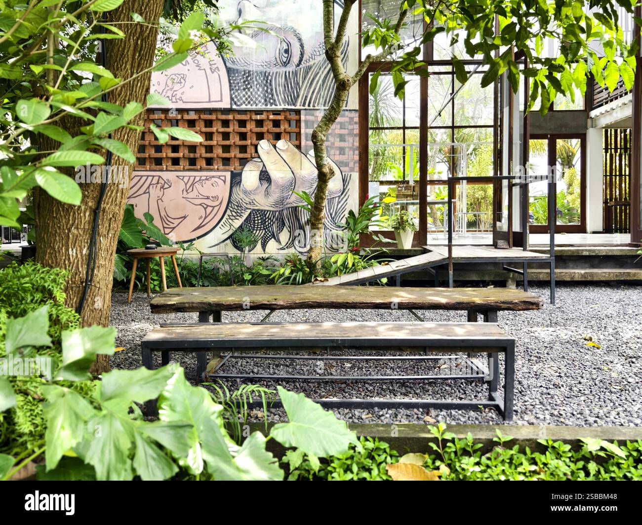 Gianyar, Bali, Indonésie : espace café extérieur avec bancs en bois, sol en gravier, verdure tropicale et murale artistique Banque D'Images