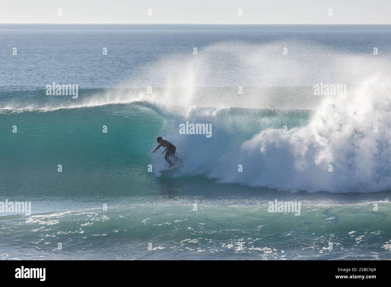 Surfeur en combinaison noire brave le surf hivernal et chevauche une vague dans les eaux bleu-vert limpides avec un jet de brise offshore Banque D'Images Surfeur en combinaison noire brave le surf hivernal et chevauche une vague dans les eaux bleu-vert limpides avec un jet de brise offshore Banque D'Images