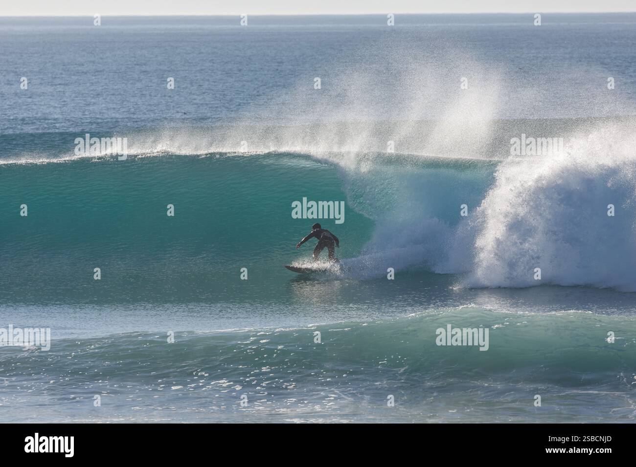 Surfeur en combinaison noire brave le surf hivernal et chevauche une vague dans les eaux bleu-vert limpides avec un jet de brise offshore Banque D'Images Surfeur en combinaison noire brave le surf hivernal et chevauche une vague dans les eaux bleu-vert limpides avec un jet de brise offshore Banque D'Images