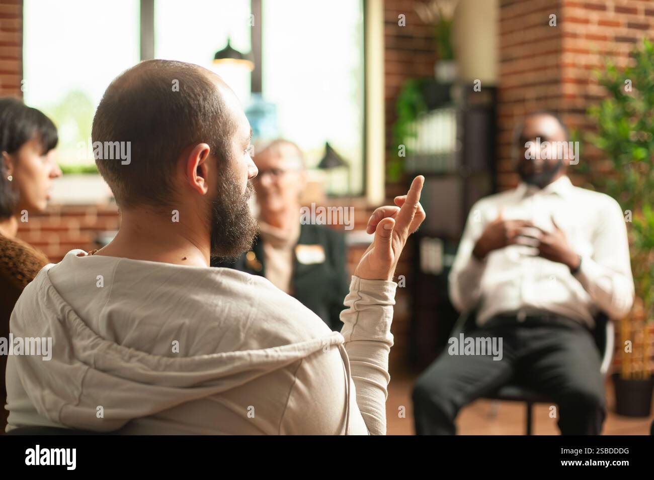 Gros plan sur un homme barbu faisant différents signes avec les doigts au cours d'une séance de thérapie de groupe. Jeune homme blanc assis en cercle et faisant des gestes de la main dans la salle de mur de briques comme les gens discutent en arrière-plan. Banque D'Images