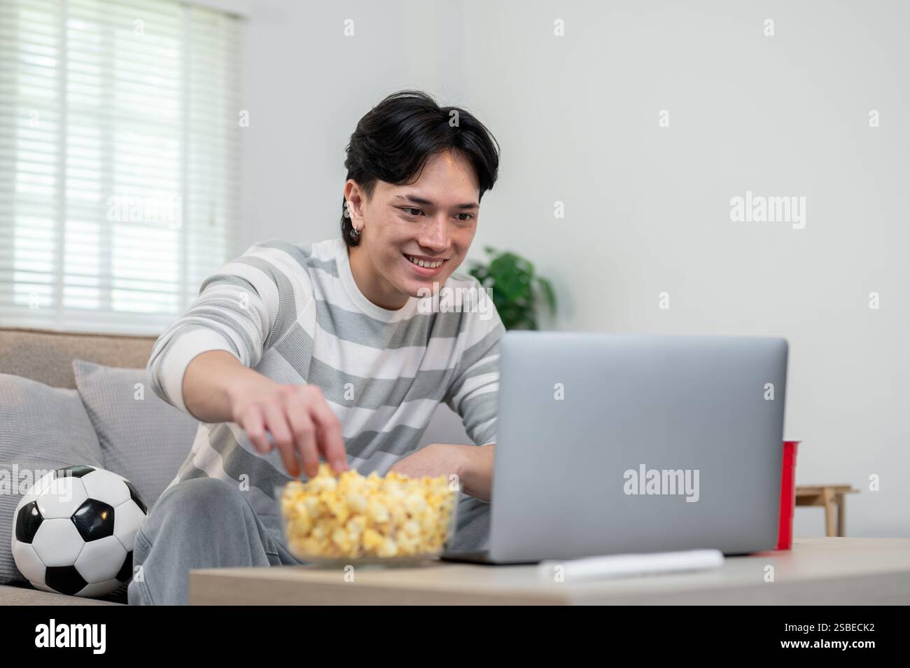 Un jeune asiatique détendu aime manger du pop-corn tout en regardant le match de son équipe de football préférée sur son ordinateur portable à la maison. art de vivre, loisirs, sport cha Banque D'Images