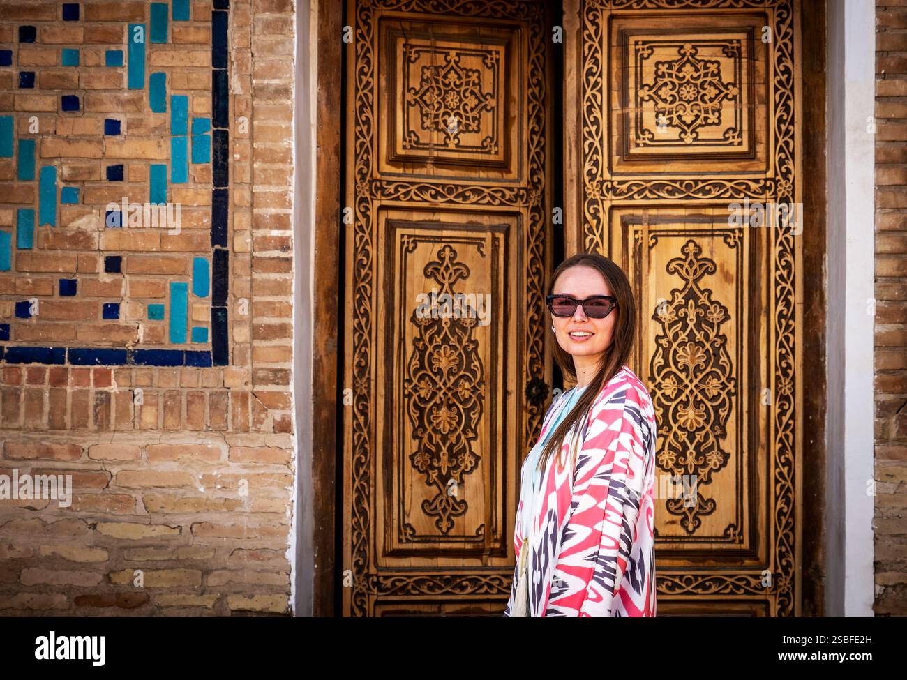 Une femme se tient devant des portes en bois au design complexe, portant des lunettes de soleil et un châle coloré à motifs. L'arrière-plan comporte un mur de briques Banque D'Images