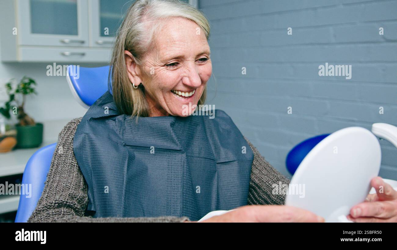 Femme mature souriant à la clinique dentaire tout en vérifiant les dents dans le miroir de main pendant le rendez-vous Banque D'Images