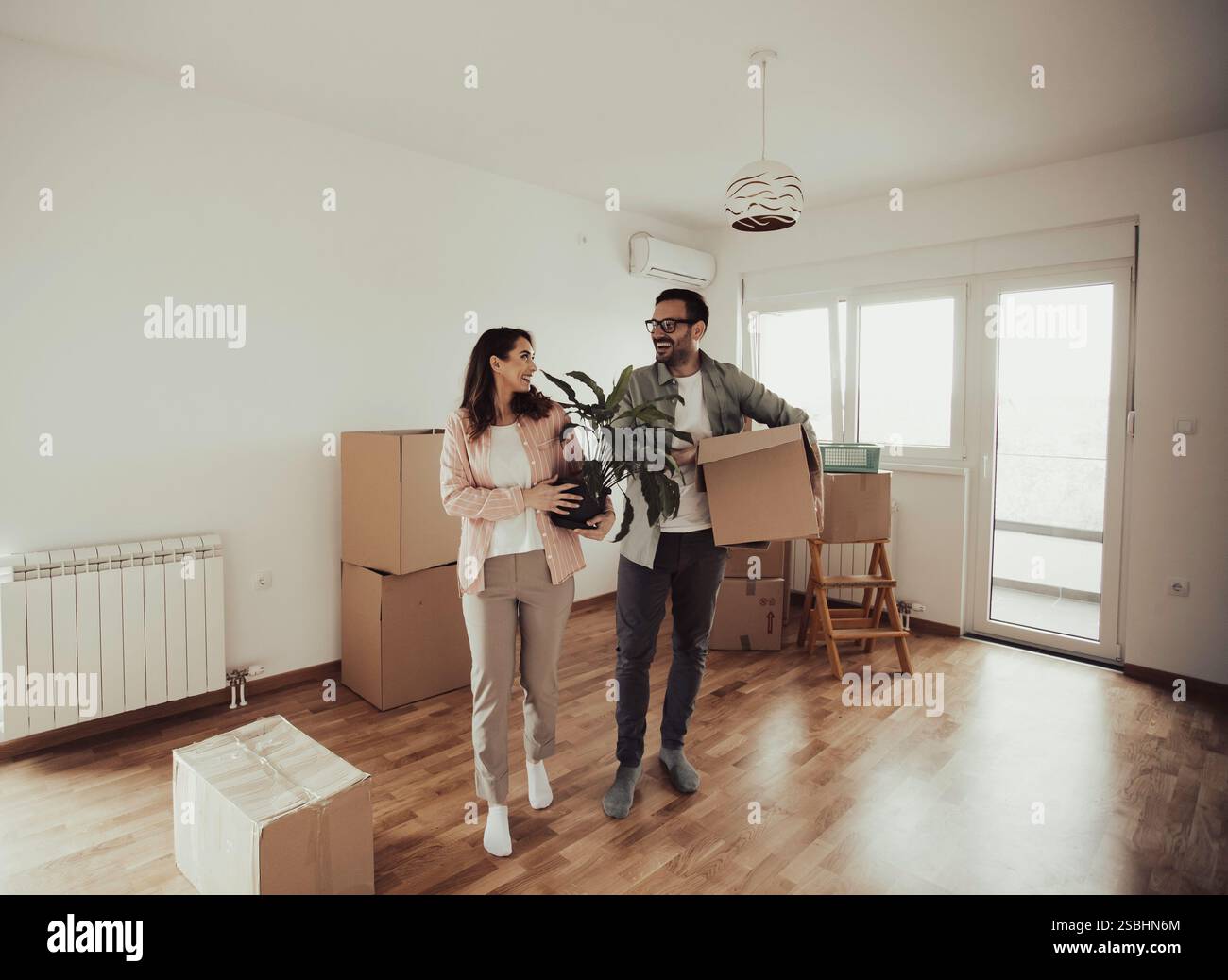 Couple heureux transportant des boîtes en carton et des plantes dans un appartement vide. Déballage des boîtes et déménagement dans une nouvelle maison ensemble Banque D'Images
