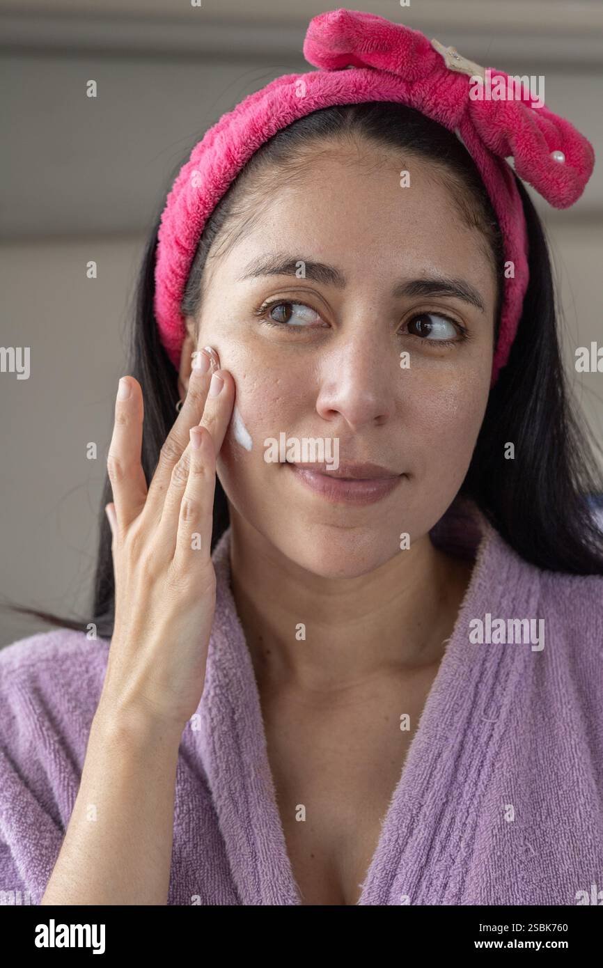 Une belle femme en peignoir et bandeau rose effectuant son rituel de nettoyage du visage. Parfait pour la beauté, les soins de la peau et le contenu d'auto-soin. Banque D'Images
