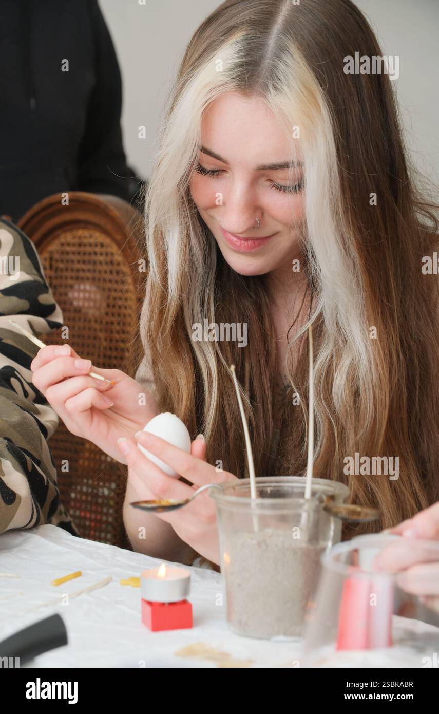 Une jeune femme aux longs cheveux bicolores décore joyeusement un œuf de Pâques en utilisant des techniques traditionnelles de cire. Elle sourit chaleureusement, immergée dans le créatif A. Banque D'Images