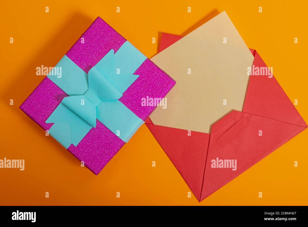 Boîte cadeau brillante colorée lumineuse décorée avec un noeud en ruban près de l'enveloppe ouverte sur fond orange vif. Parfait pour les célébrations, les cadeaux ou les événements Banque D'Images