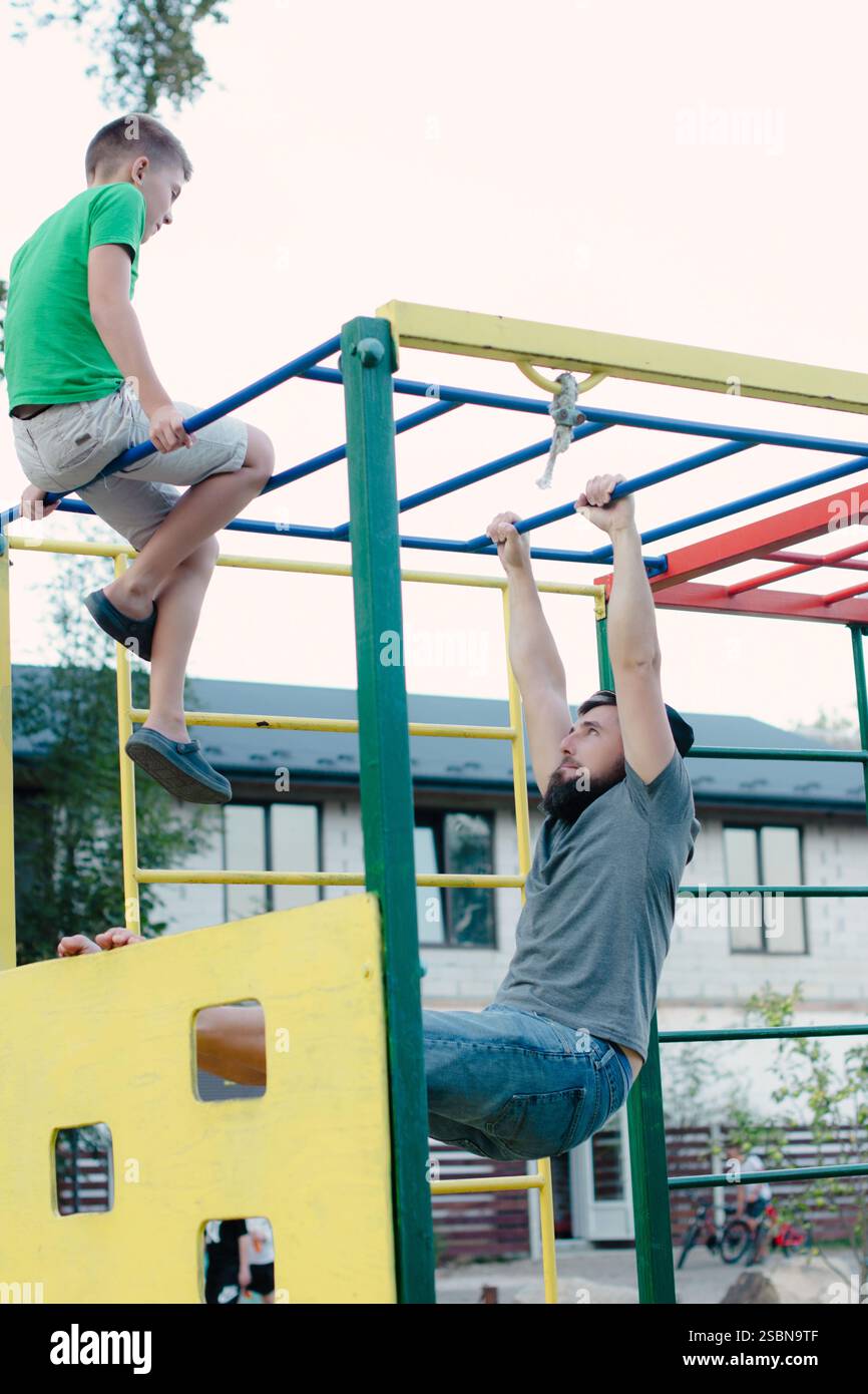 Un père et son fils jouent sur un cadre d'escalade extérieur. Le père est suspendu à la barre supérieure, tandis que son fils est assis sur une barre inférieure, les deux regardant Banque D'Images