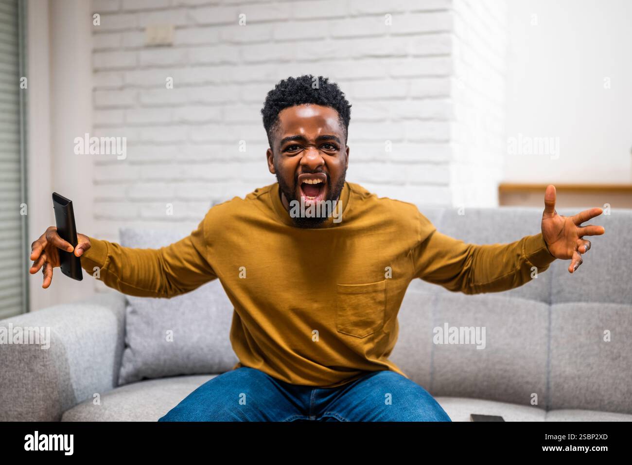 Jeune homme afro-américain déçu criant et faisant des gestes de colère tout en regardant un match de football à la télévision à son domicile. Banque D'Images