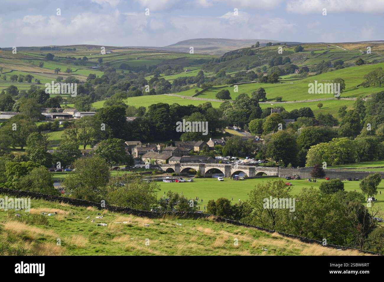 Burnsall niché dans la vallée (pont voûté, maisons et chalets, champs verdoyants à flanc de colline, vue longue distance, été) - Yorkshire Dales, Angleterre, Royaume-Uni. Banque D'Images