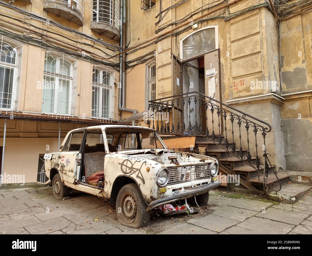 Vieille voiture délabrée dans la cour en face de l'ancien bâtiment avec des escaliers ; pneus crevés ; partiellement démontés, démontés ; concept de pourriture - Image de stock capturée avec un smartphone