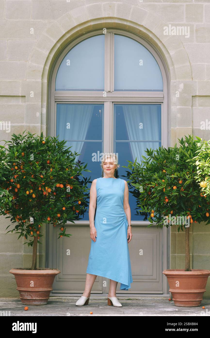 Portrait complet de la longueur du corps de belle femme d'âge moyen de 50 -55 ans, portant une robe bleue, posant dehors à côté d'orangers plantés Banque D'Images