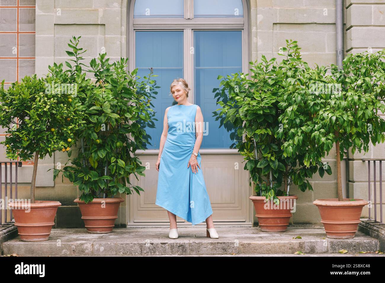 Portrait complet de la longueur du corps de belle femme d'âge moyen de 50 -55 ans, portant une robe bleue, posant dehors à côté de citronniers plantés Banque D'Images