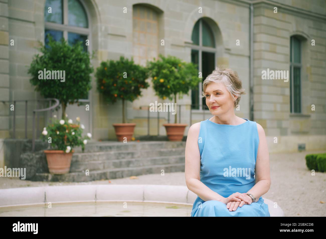 Portrait en plein air de belle femme mature de 50 - 55 ans, portant une robe bleue, se relaxant à côté d'une fontaine dans un jardin confortable, va méditerranéenne d'été Banque D'Images