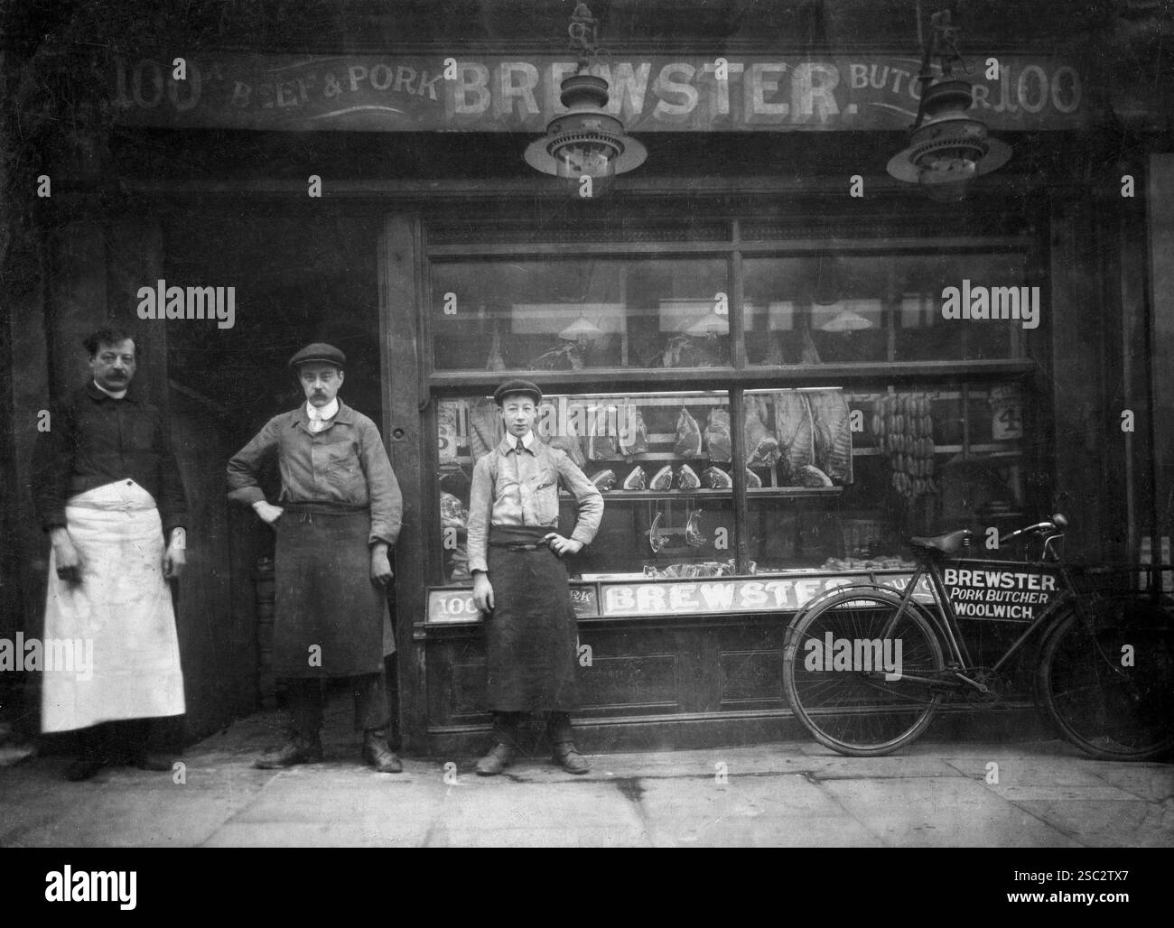 Londres, Angleterre .c.1910 – Une photographie de la boucherie de bœuf et de porc Brewster, située au 100A Woolwich High Street, au sud-est de Londres. L'entreprise a été fondée par Albert George Brewster (1868-1929), qui a également dirigé une deuxième boutique à Hare Street, Woolwich. La photographie montre Albert Brewster avec deux membres de son personnel. Un vélo de livraison se tient devant la vitrine du magasin. Après la mort d’Albert, l’entreprise fut poursuivie par sa femme, Gertrude, jusqu’à sa mort en 1944. Banque D'Images