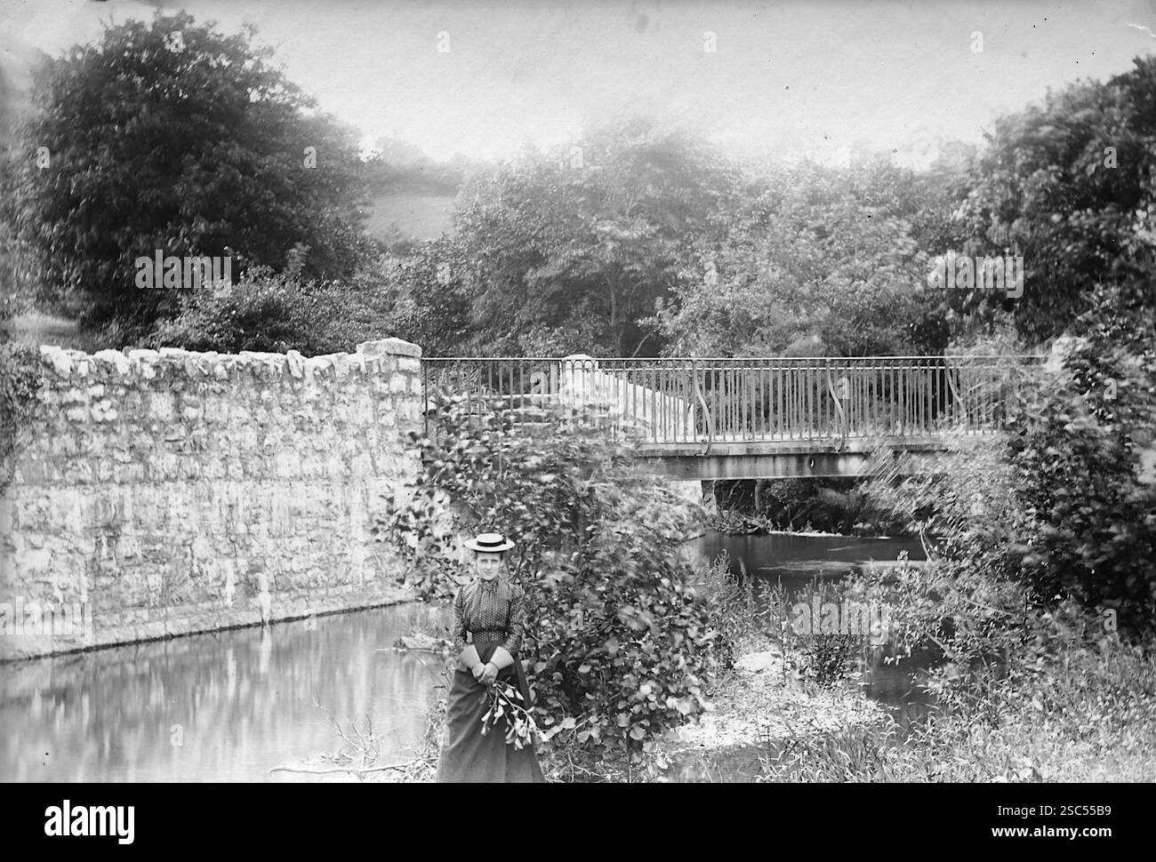 Une dame, avec des fleurs, dans un paysage fluvial avec un petit pont en arrière-plan, River (Afon) Conwy, Nord du pays de Galles. C1900s, monochrome : d'une importante collection historique de photographies originales et inattribuées de l'albumen de la fin de l'époque victorienne au début de l'époque édouardienne : une tournée britannique comprenant le Staffordshire, le Warwickshire et le nord du pays de Galles. La qualité des originaux était variable et la plupart étaient autour de 108x165mm. Banque D'Images