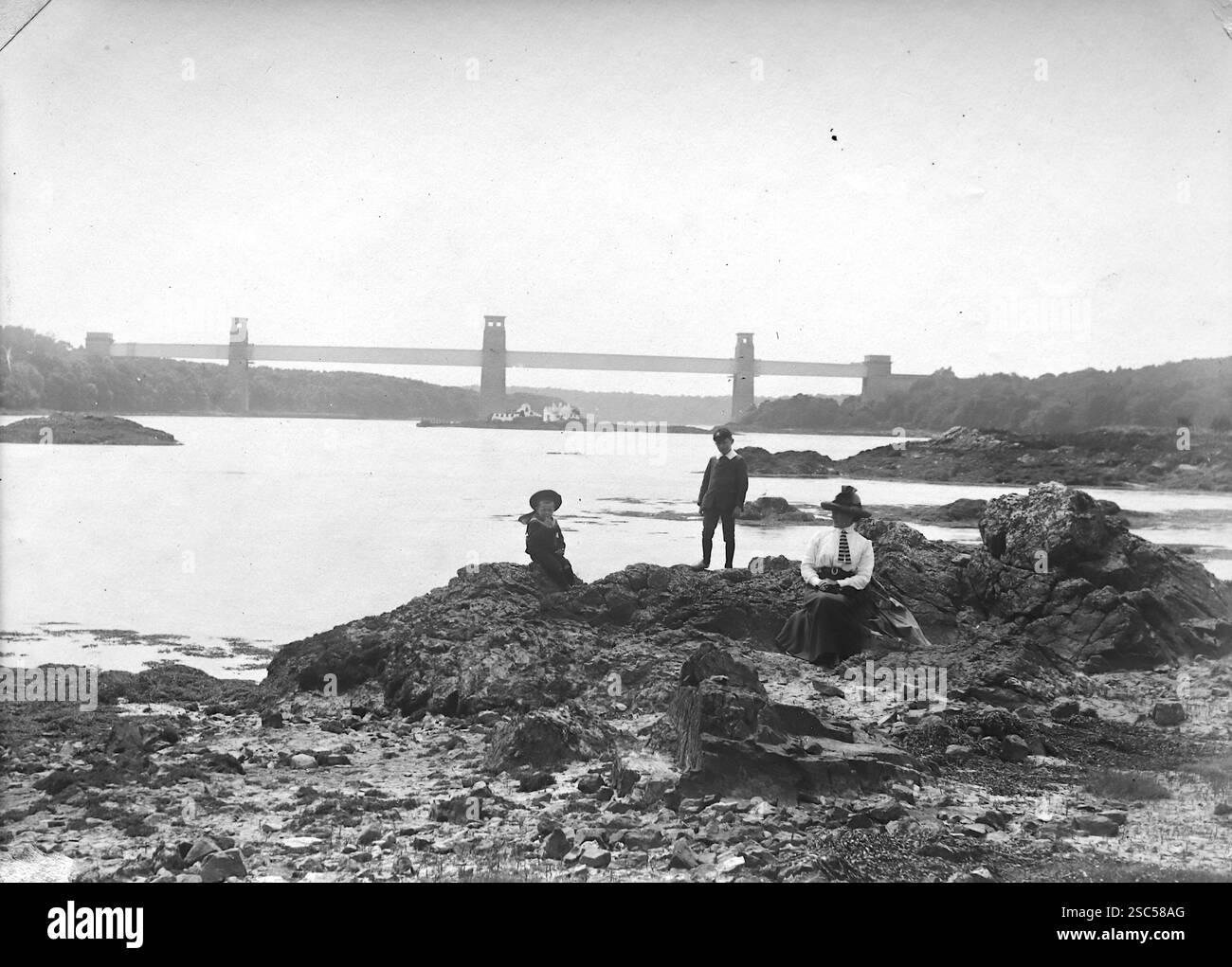 Une dame et deux enfants sur le rivage. À l'arrière-plan se trouve le pont tubulaire (maintenant connu sous le nom de pont Britannia) qui traverse le détroit de Menai (ou Afon Menai en gallois), une étroite étendue d'eau qui sépare l'île d'Anglesey du continent de Gwynedd dans le nord du pays de Galles. C1900s, monochrome : d'une importante collection historique de photographies originales et inattribuées de l'albumen de la fin de l'époque victorienne au début de l'époque édouardienne : une tournée britannique comprenant le Staffordshire, le Warwickshire et le nord du pays de Galles. La qualité des originaux était variable et la plupart étaient autour de 108x165mm. Banque D'Images