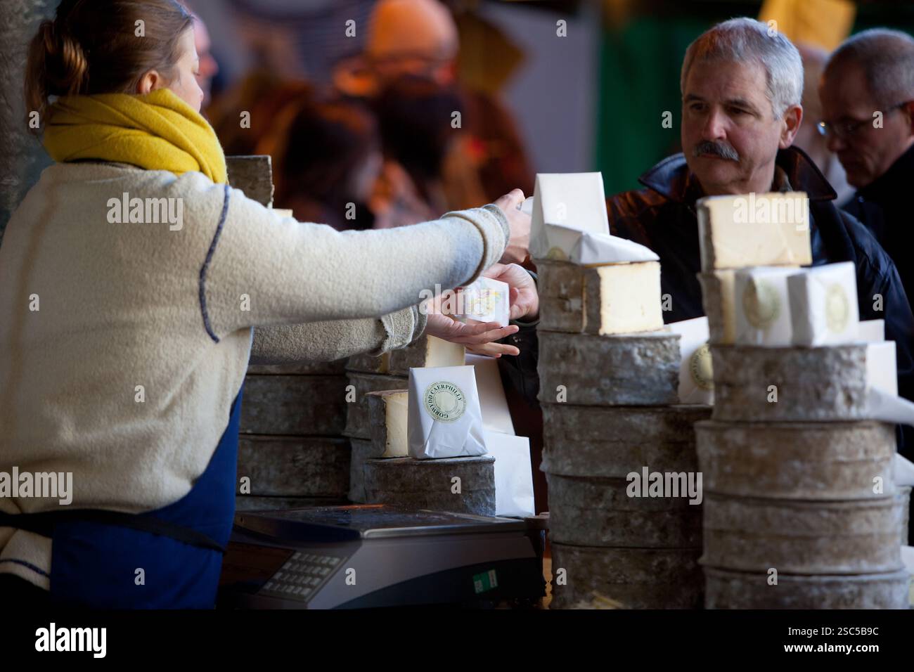 Un client achète du fromage à un étal de marché extérieur, avec des meules de fromage empilées visibles. Banque D'Images