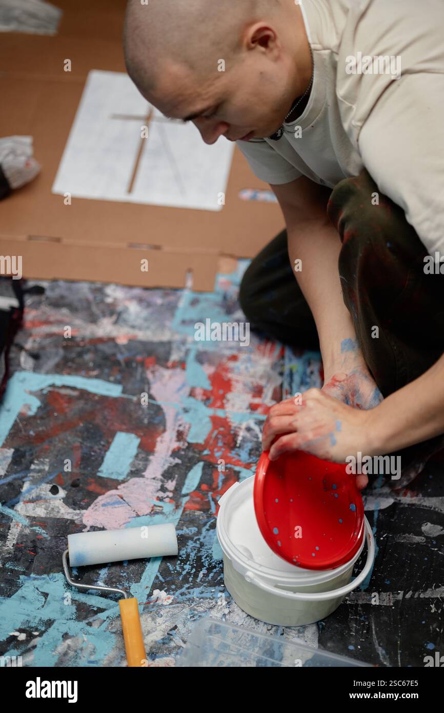 Plan vertical de l'artiste masculin ouvrant seau en plastique avec impression croisée de dessin de peinture blanche sur les vêtements à l'aide d'un pochoir en papier sur le sol de l'atelier teinté Banque D'Images