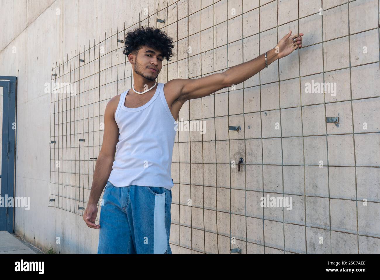 Un beau jeune homme latino avec un style personnel tenant une clôture d'un grand mur de béton. Banque D'Images