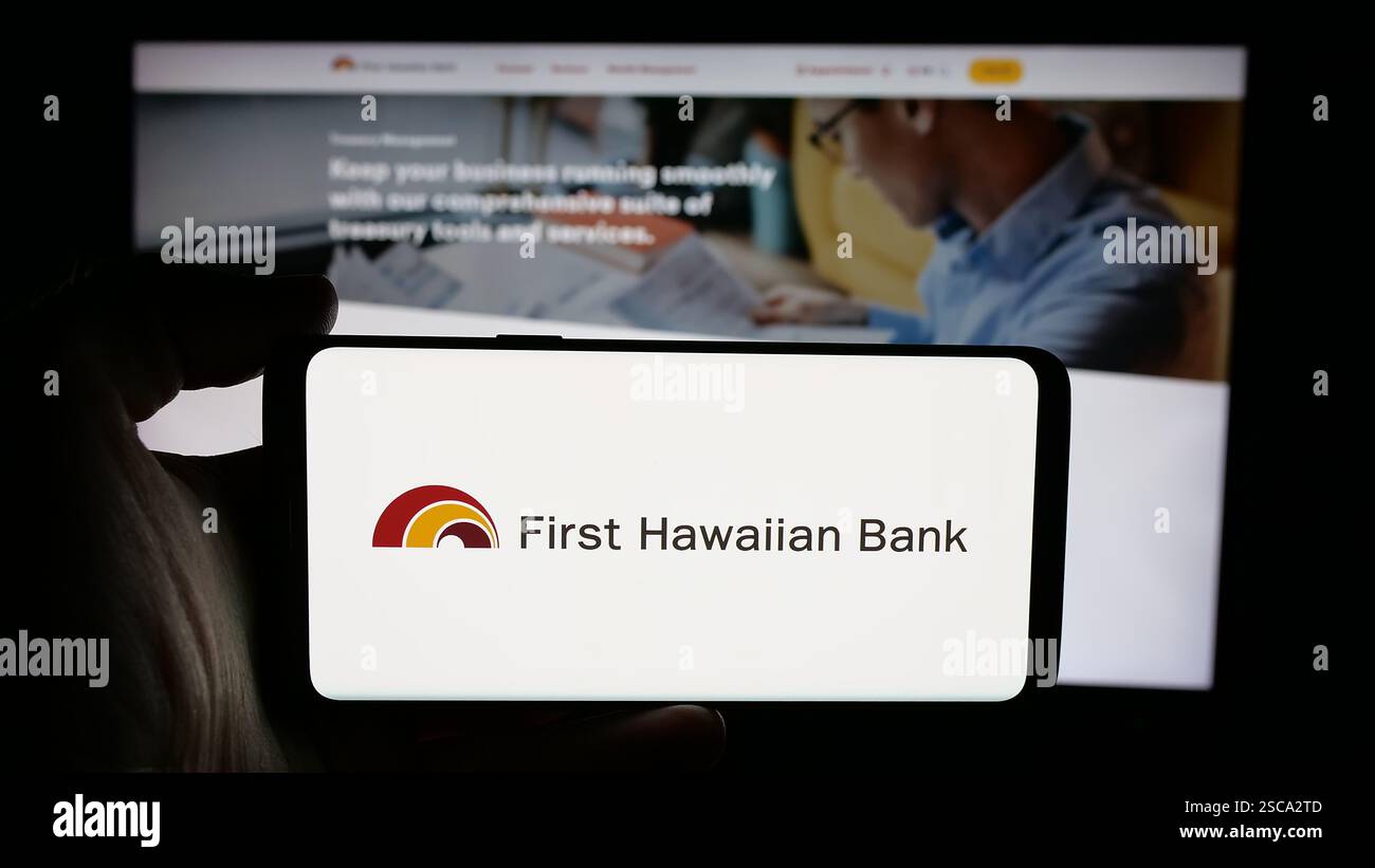 Dans cette illustration photo, une personne tient un téléphone portable avec le logo de la société de services financiers américaine First Hawaiian Inc sur l'écran devant la page Web. Banque D'Images