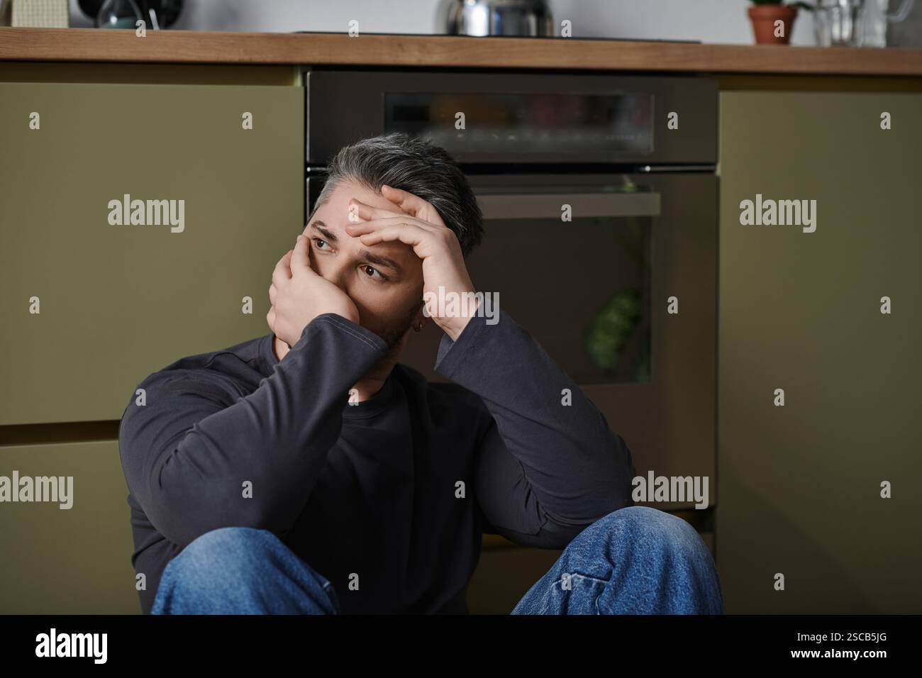 Un bel homme est assis sur le sol de la cuisine, perdu dans la pensée profonde. Banque D'Images