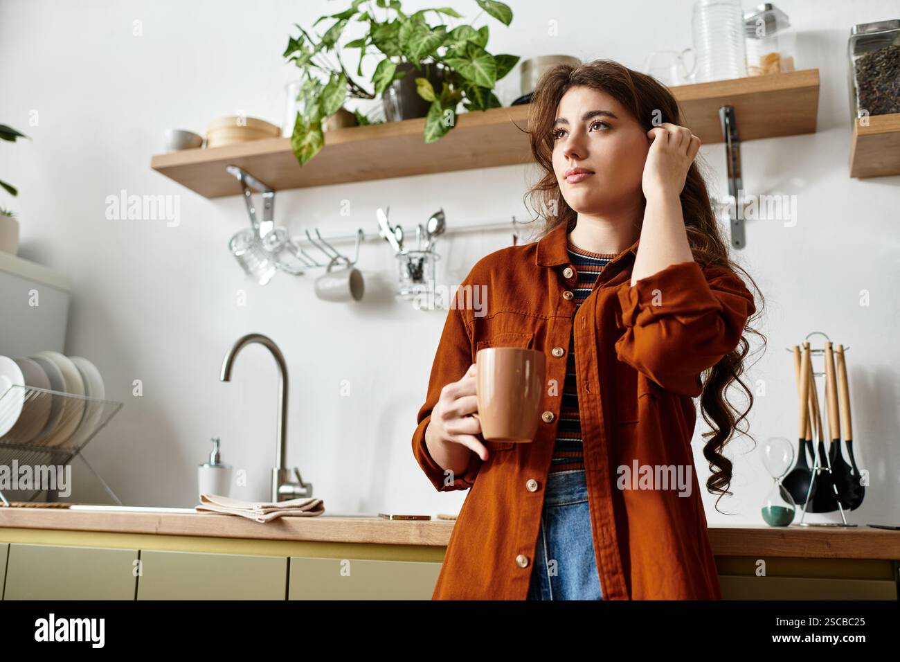 Une femme se tient réfléchie dans une cuisine lumineuse, tenant une tasse et perdue dans la contemplation profonde. Banque D'Images