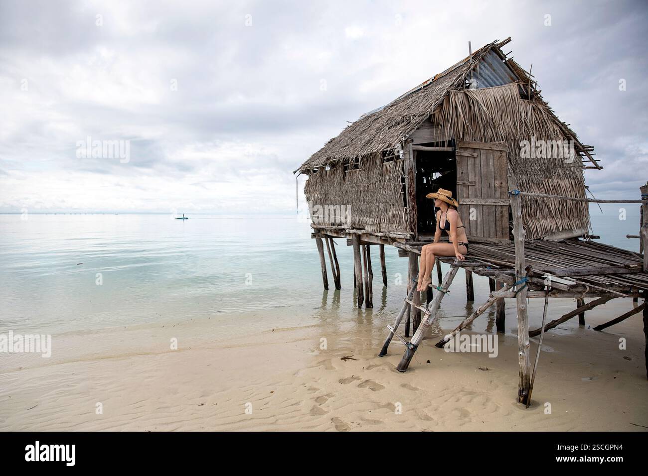 Femme en maillot de bain noir et un chapeau de paille assis sur l'échelle des maisons traditionnelles en bois du peuple Bajau sur une partie éloignée de Sulawesi, Indonésie Banque D'Images