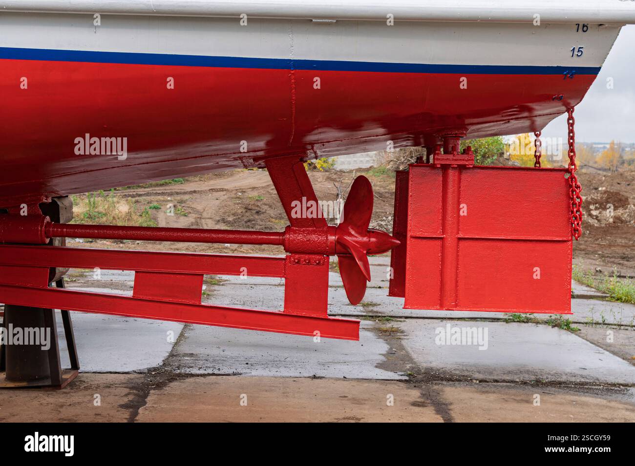 Bateau en cale sèche avec une coque rouge et blanche, montrant son système de gouvernail et son hélice, soutenus par des blocs de bois et de métal, en vue rapprochée du chantier naval Banque D'Images