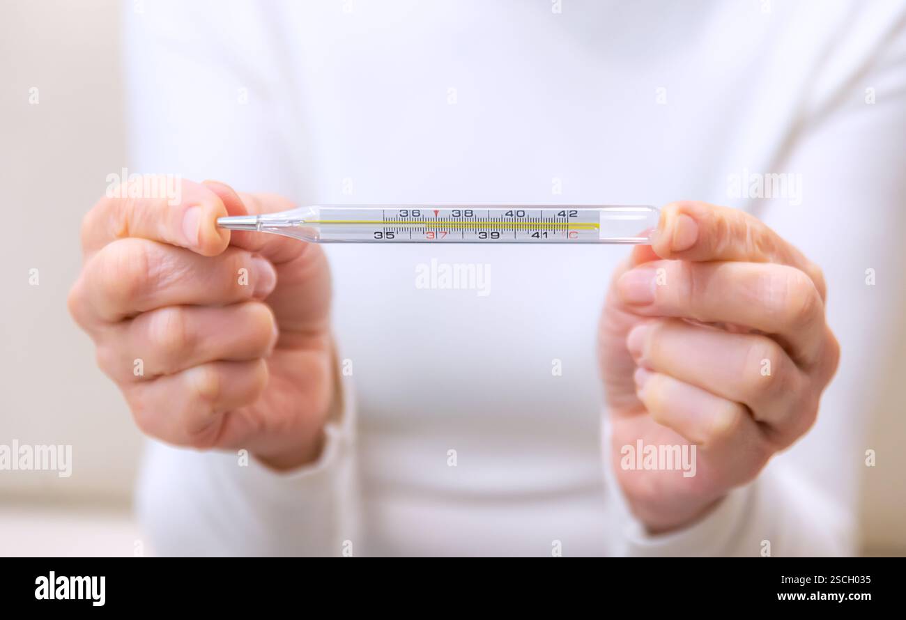 La personne tient un thermomètre pour mesurer la température corporelle, reflétant une préoccupation pour la santé et le bien-être dans un milieu domestique. Banque D'Images
