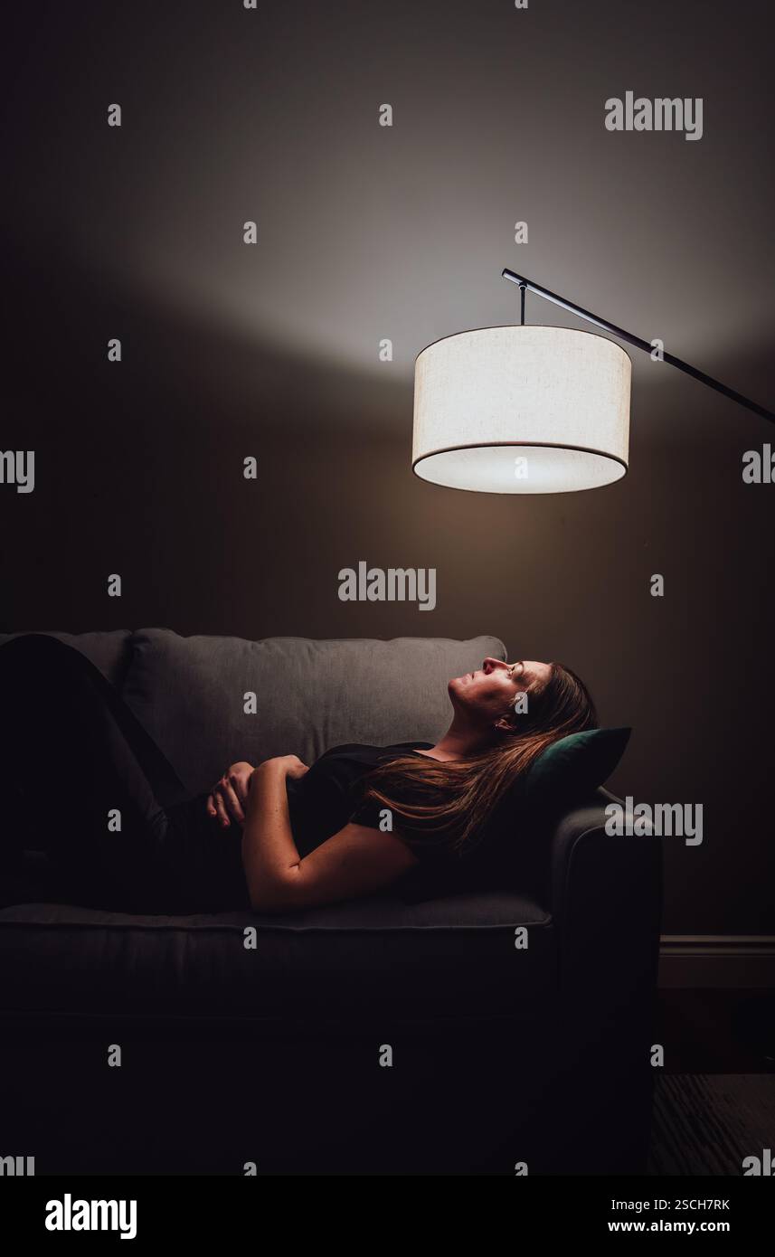 Femme allongée sur un canapé dans une pièce sombre regardant vers le haut à la lumière de la lampe. Banque D'Images