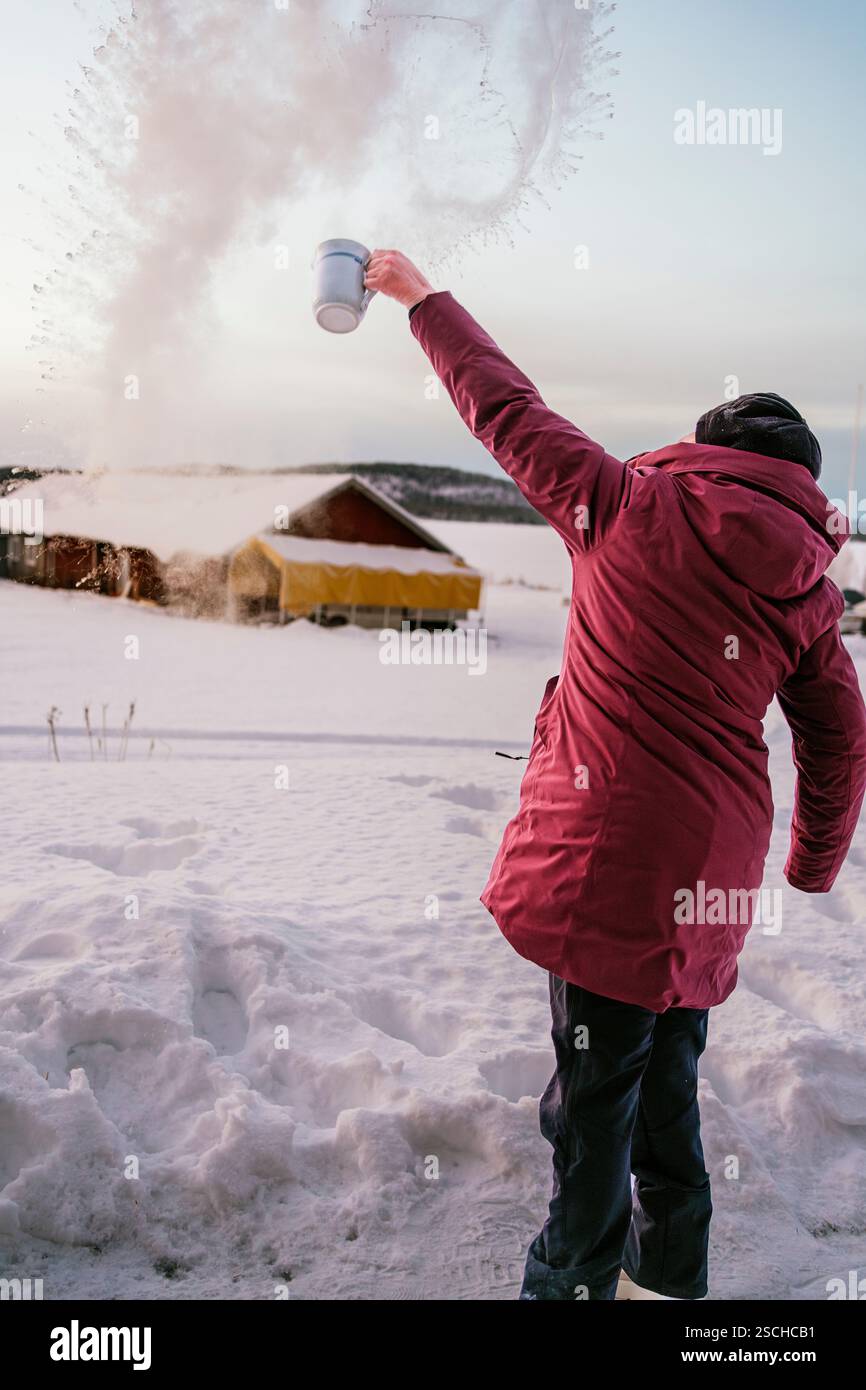 Une personne au manteau rouge jette de l'eau dans l'air glacial de la Laponie suédoise, créant un jet impressionnant de cristaux de glace contre le dos enneigé Banque D'Images