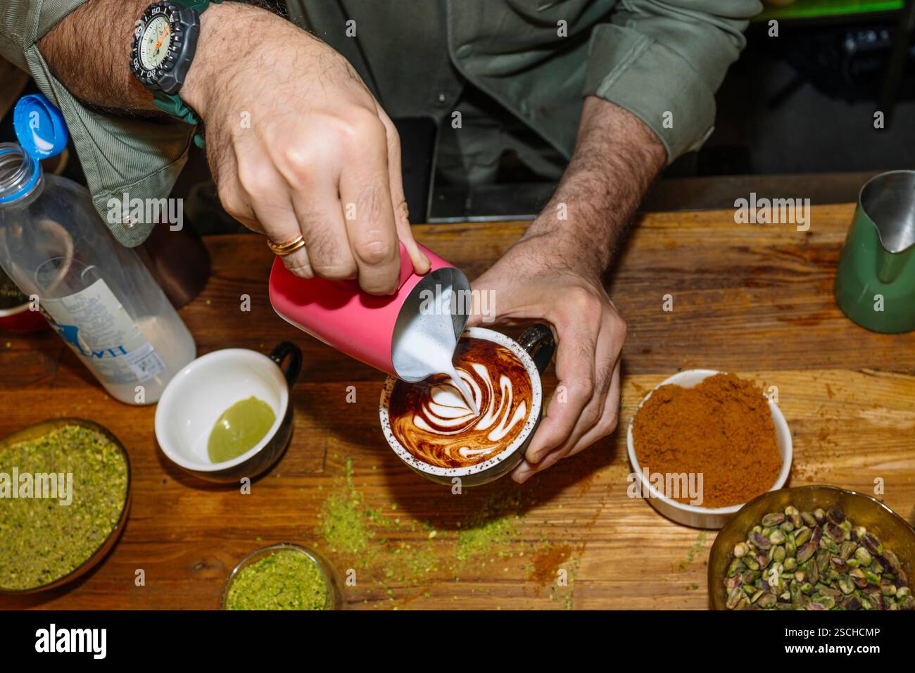 Un barista confectionne un latte art complexe sur un cappuccino crémeux, entouré d'ingrédients comme la crème, la pistache et les épices, mettant en valeur le talent artistique Banque D'Images