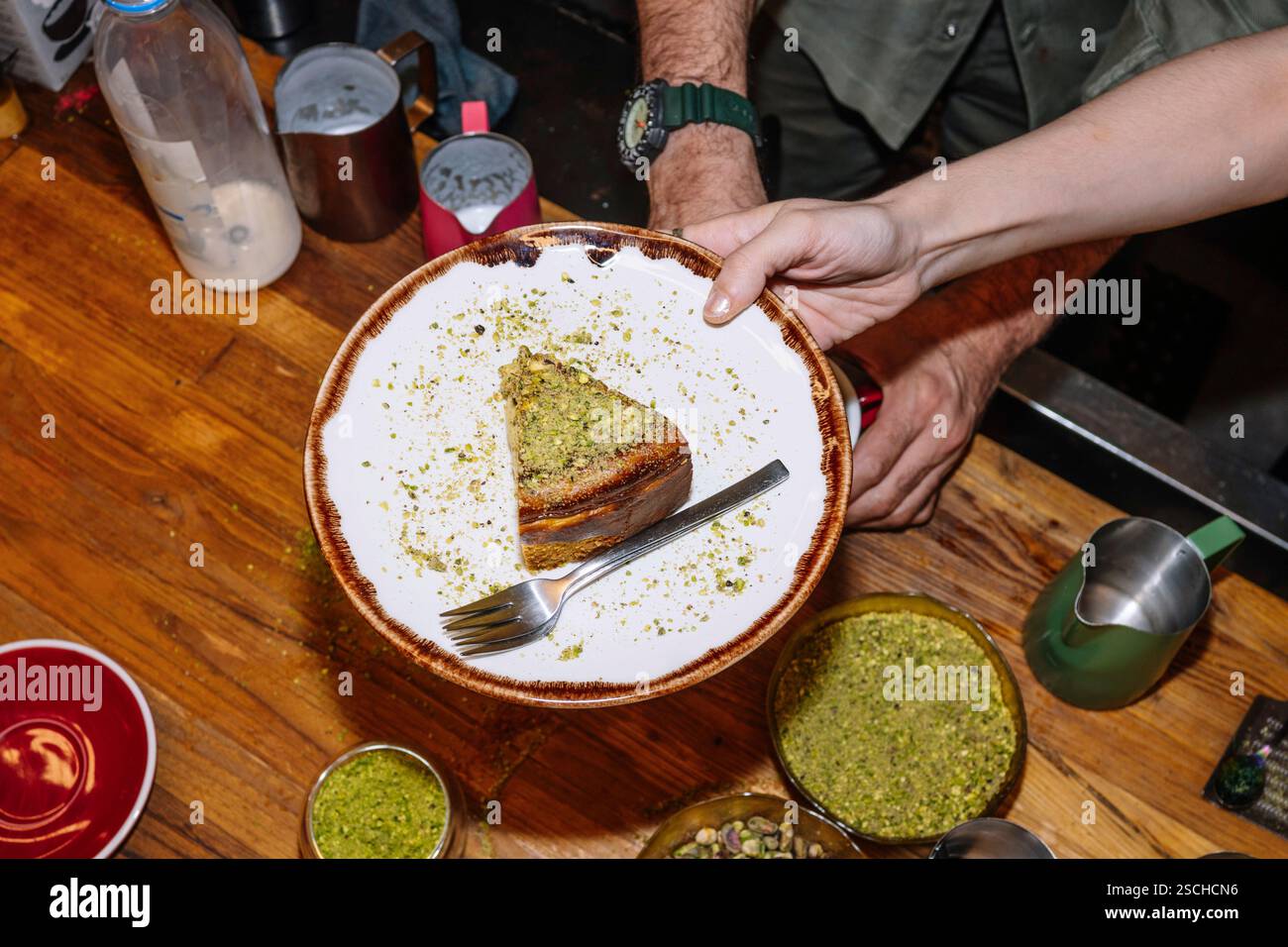 Une tranche plaquée de cheesecake surmontée de pistaches écrasées est présentée sur une assiette rustique avec une fourchette. L'arrière-plan montre un kitche de café occupé Banque D'Images